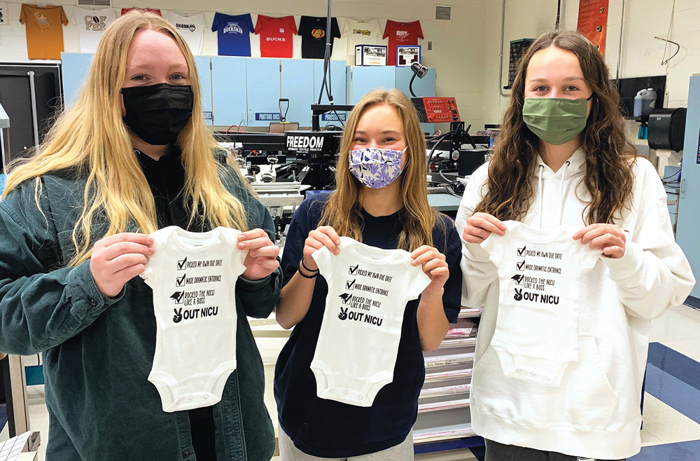 Three female high school students wearing facial masks hold up white onesies on which is printed the following: “Picked my own due date. Made dramatic entrance. Rocked the NICU like a boss. Out NICU.” 